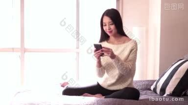 美丽的亚洲女人在家里的床上用手机发<strong>短信</strong>，微笑着